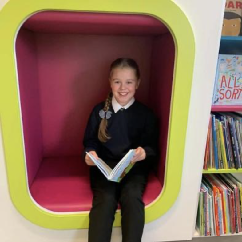 A girl is sat smiling whilst reading a book in the school library